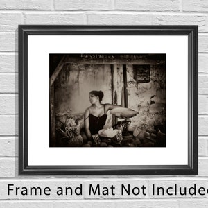 May include: A black and white photograph of a woman standing in a doorway with a scale in front of her. The woman is wearing a dark dress and has her hair pulled back. The scale is old and has a wooden base. The photo is taken in a dimly lit room and has a vintage feel.