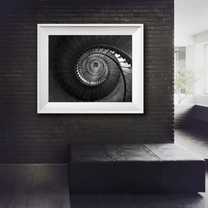 May include: A black and white photograph of a spiral staircase framed in a white frame. The photograph is hanging on a black brick wall.