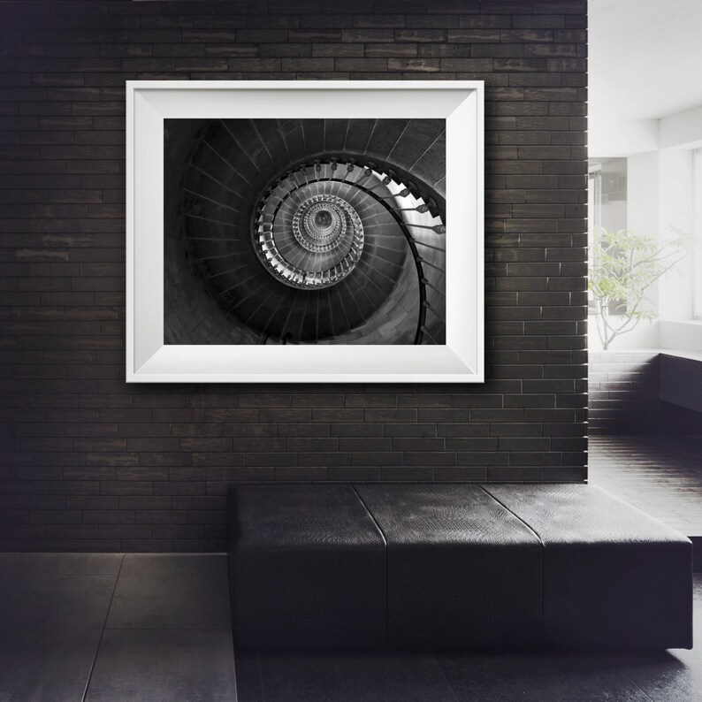 May include: A black and white photograph of a spiral staircase framed in a white frame. The photograph is hanging on a black brick wall.