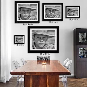 May include: Black and white framed portrait prints of a young person wearing a straw hat. The prints are displayed in various sizes, including 30 x 24 inches, 24 x 20 inches, 20 x 16 inches, 14 x 11 inches, and 10 x 8 inches. The portraits are hung on a white wall above a wooden table.