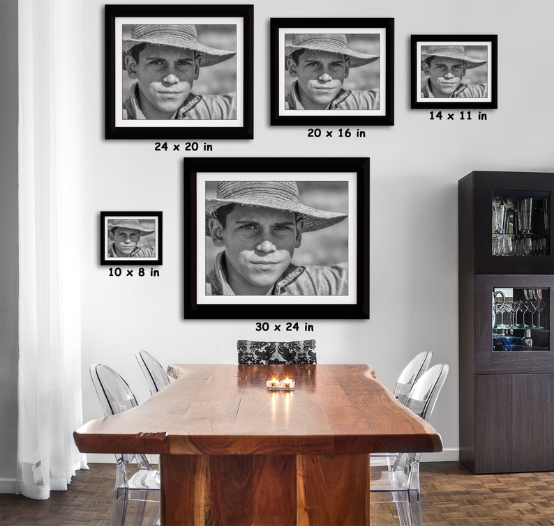 May include: Black and white framed portrait prints of a young person wearing a straw hat. The prints are displayed in various sizes, including 30 x 24 inches, 24 x 20 inches, 20 x 16 inches, 14 x 11 inches, and 10 x 8 inches. The portraits are hung on a white wall above a wooden table.