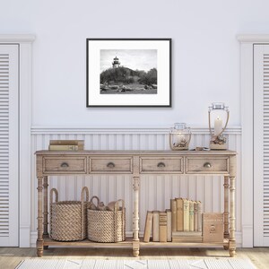 May include: A black and white photograph of a lighthouse on a rocky island. The photo is framed in a black frame and is hanging on a white wall above a wooden console table. The console table has two drawers and is decorated with two wicker baskets, a candle lantern, and a collection of books.