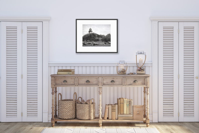 May include: A black and white photograph of a lighthouse on a rocky island. The photo is framed in a black frame and is hanging on a white wall above a wooden console table. The console table has two drawers and is decorated with two wicker baskets, a candle lantern, and a collection of books.