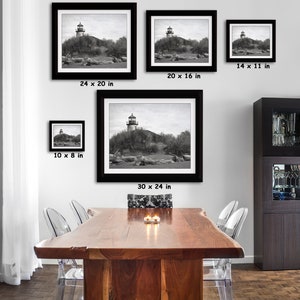 May include: A black and white photograph of a lighthouse on a small island. The lighthouse is tall and slender, with a white tower and a black roof. The island is covered in trees and bushes. The photo is framed in a black frame and is available in various sizes: 10 x 8 inches, 14 x 11 inches, 20 x 16 inches, 24 x 20 inches, and 30 x 24 inches.