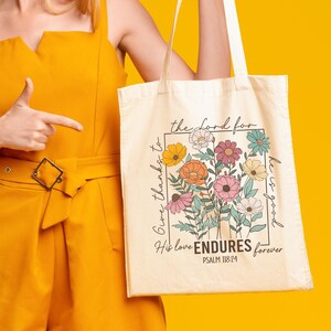May include: A woman wearing a straw hat and sunglasses is holding a white tote bag with a floral design and the text "Give thanks to the Lord for His love ENDURES forever Psalm 136:14".