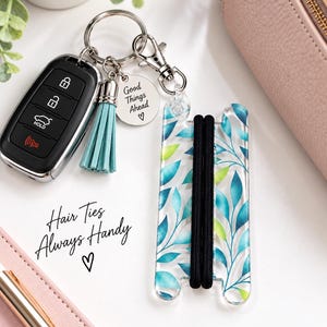 May include: A keychain featuring a black car key fob, a light blue tassel, and a round silver charm engraved with the text Good Things Ahead and a heart. Attached is a clear acrylic holder with a blue and green leaf pattern holding two black hair ties.