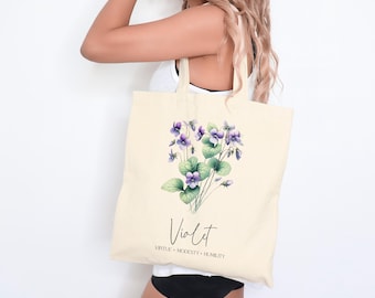 May include: A beige tote bag with a floral design featuring purple and green violets. The text "Violet" is printed below the flowers, along with the words "Virtue, Modesty, Humility".