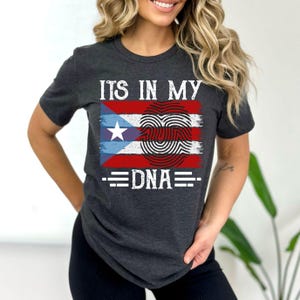 Puede incluir: Una camiseta gris oscuro con una impresión blanca que dice "ITS IN MY DNA" con un diseño de bandera puertorriqueña roja, blanca y azul con una huella digital en el medio.