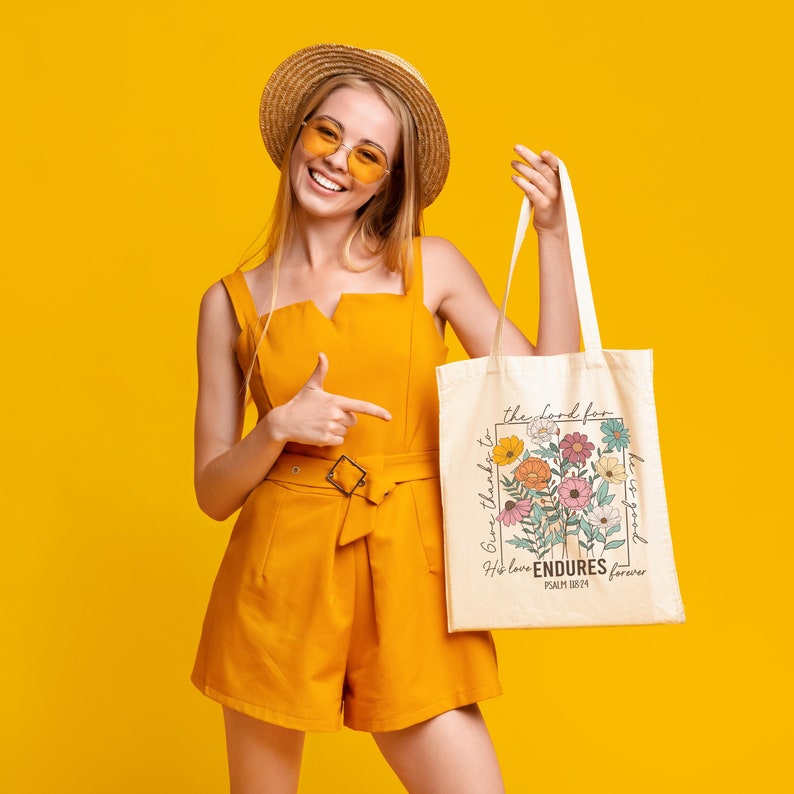 May include: A woman wearing a straw hat and sunglasses is holding a white tote bag with a floral design and the text "Give thanks to the Lord for His love ENDURES forever Psalm 136:14".