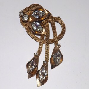 May include: Gold-toned vintage brooch with a woven mesh design and dangling teardrop-shaped pendants. The brooch features clear, faceted crystal accents. A classic piece of jewelry.