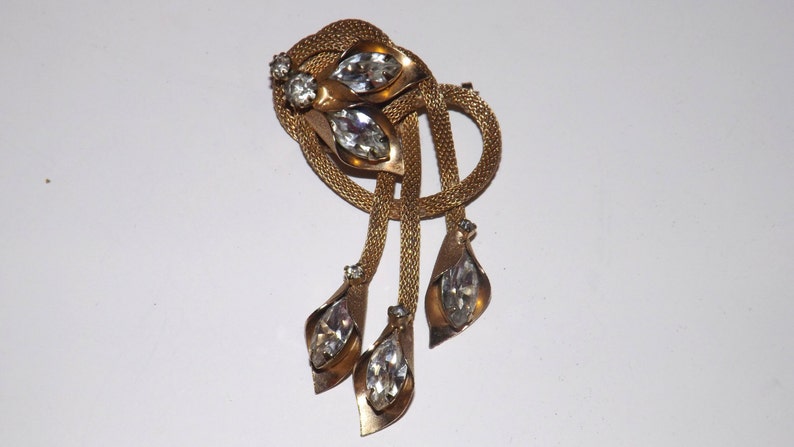 May include: Gold-toned vintage brooch with a woven mesh design and dangling teardrop-shaped pendants. The brooch features clear, faceted crystal accents. A classic piece of jewelry.