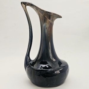 May include: A dark blue ceramic vase with a long, curved handle. The vase has a bulbous base and a flared opening. The handle curves gracefully from the base to the top. The vase has a glossy finish with hints of brown and gray.