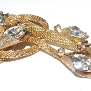 May include: A vintage gold-tone brooch featuring a woven mesh design with three clear, faceted crystal accents. The brooch has a leaf-like design with large, clear crystals and smaller accent stones.