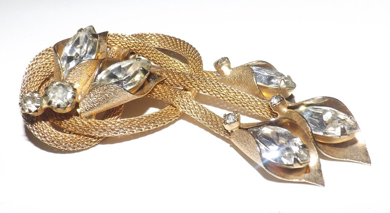 May include: A vintage gold-tone brooch featuring a woven mesh design with three clear, faceted crystal accents. The brooch has a leaf-like design with large, clear crystals and smaller accent stones.