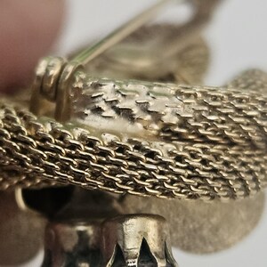 May include: Close-up of a gold-toned brooch with a textured chain design. The brooch features a decorative element with a clear, faceted stone. The pin and clasp are visible, suggesting it's a vintage or antique piece.