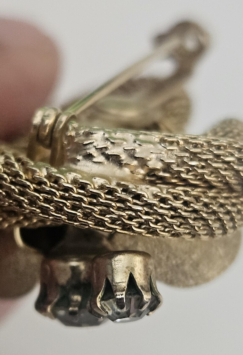May include: Close-up of a gold-toned brooch with a textured chain design. The brooch features a decorative element with a clear, faceted stone. The pin and clasp are visible, suggesting it's a vintage or antique piece.