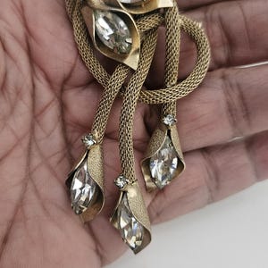 May include: Vintage gold-tone brooch with a floral design. The brooch features three dangling pendants with teardrop-shaped clear crystals. The brooch has a mesh-like chain and is set with clear rhinestones.