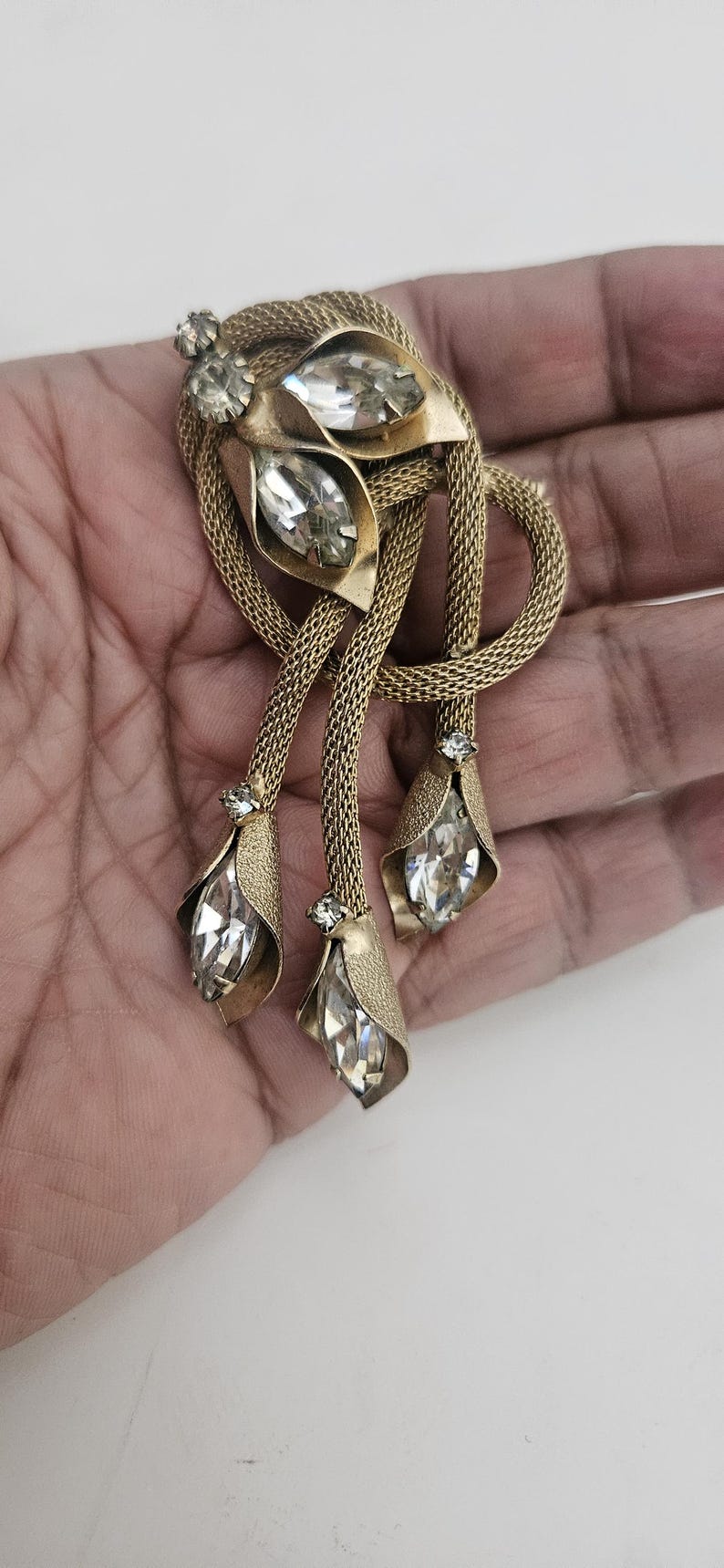 May include: Vintage gold-tone brooch with a floral design. The brooch features three dangling pendants with teardrop-shaped clear crystals. The brooch has a mesh-like chain and is set with clear rhinestones.
