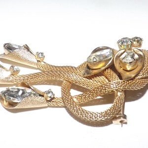 May include: A gold-toned vintage brooch with a woven design and clear rhinestone accents. The brooch features three fan-shaped elements and a central knot design, with multiple clear rhinestones.