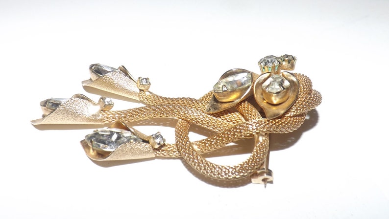 May include: A gold-toned vintage brooch with a woven design and clear rhinestone accents. The brooch features three fan-shaped elements and a central knot design, with multiple clear rhinestones.