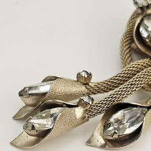 May include: Close-up of a vintage gold-toned brooch with clear, faceted crystal accents. The brooch features a textured leaf design with three large crystals and a woven chain detail. The crystals are set in a prong setting.