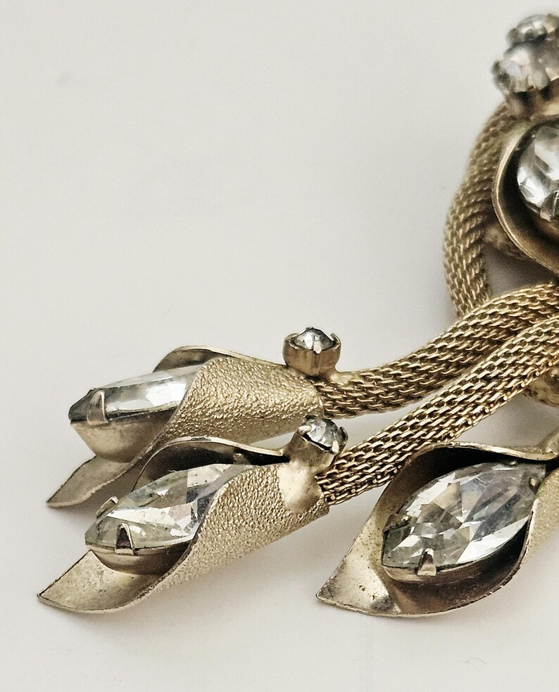 May include: Close-up of a vintage gold-toned brooch with clear, faceted crystal accents. The brooch features a textured leaf design with three large crystals and a woven chain detail. The crystals are set in a prong setting.