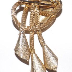 May include: A gold-toned brooch with a textured, woven design. The brooch features a knotted top section with three dangling, cone-shaped pendants. The pin closure is visible on the back, indicating it's designed to be worn on clothing.