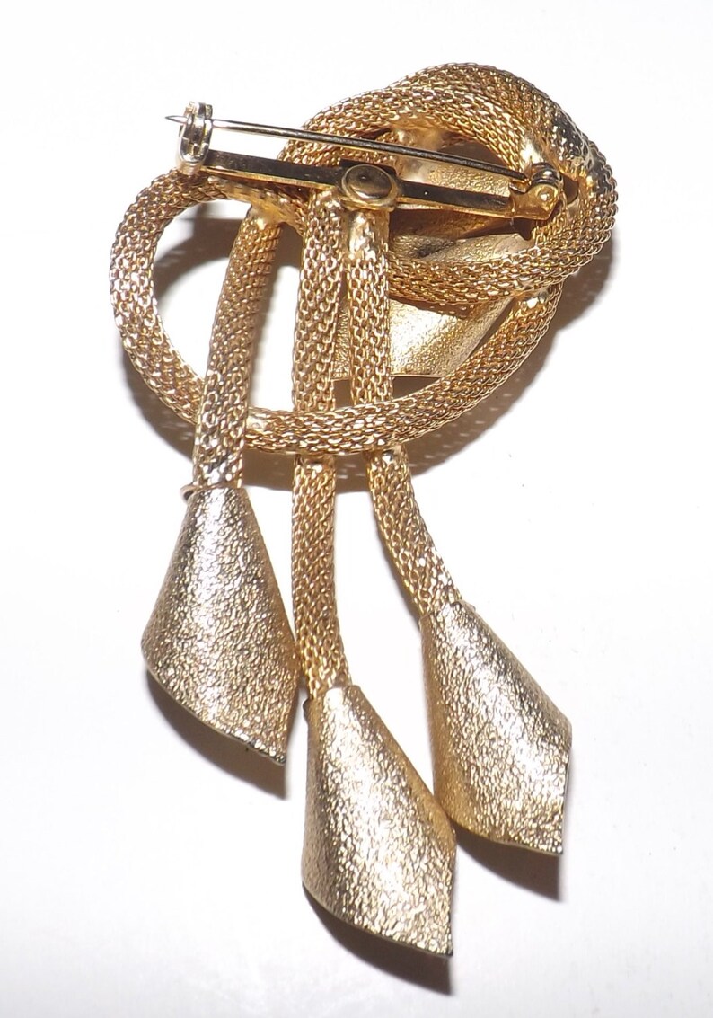 May include: A gold-toned brooch with a textured, woven design. The brooch features a knotted top section with three dangling, cone-shaped pendants. The pin closure is visible on the back, indicating it's designed to be worn on clothing.