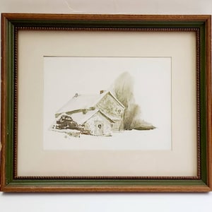 May include: A framed watercolor painting of a small, stone house with a snow-covered roof. The house has a door with a wreath and a window. The house is surrounded by trees and snow.