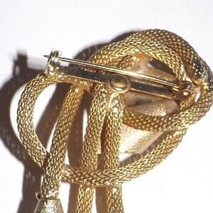 May include: A gold-toned brooch with a textured, rope-like design. The brooch features a looped knot with dangling elements and a pin closure on the back. The overall design is intricate and detailed, with a vintage aesthetic.