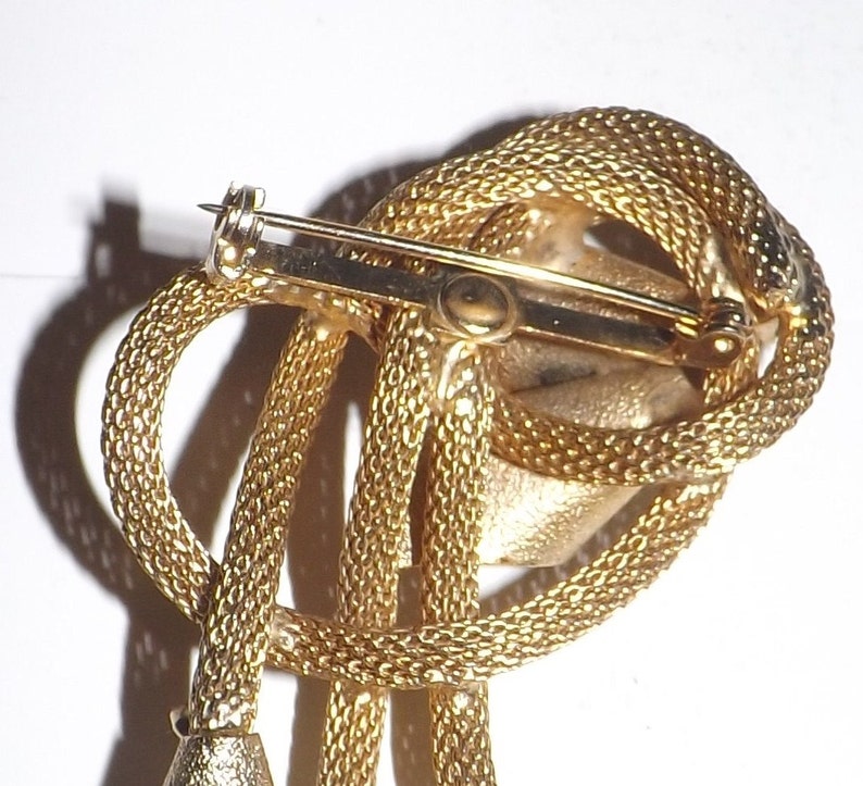 May include: A gold-toned brooch with a textured, rope-like design. The brooch features a looped knot with dangling elements and a pin closure on the back. The overall design is intricate and detailed, with a vintage aesthetic.