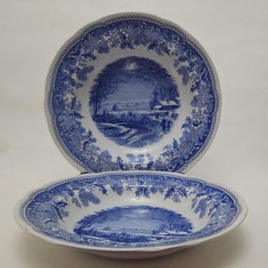 May include: Two blue and white ceramic bowls with a winter landscape scene. The bowls have a scalloped edge and a decorative border. The scene depicts a bridge, trees, and a house. The bowls are likely for serving soup or cereal.
