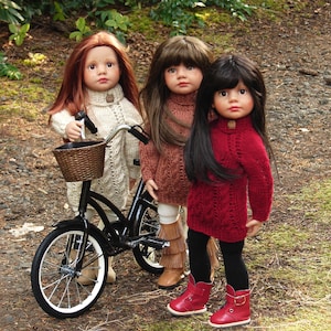 Peut inclure: Trois poupées portant des pulls et des bottes se tiennent dans un décor de forêt. Une poupée tient un vélo noir avec un panier brun. Les poupées portent des pulls bruns, rouges et crème.