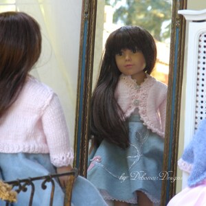 May include: A doll wearing a pink cardigan and a blue skirt with white lace trim. The doll is standing in front of a mirror and looking at herself. The mirror is framed in wood with a blue trim. The text "by Debonair Designs" is visible in the bottom right corner of the image.