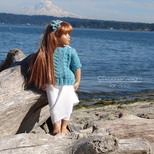 May include: A doll wearing a blue knitted sweater and a white skirt stands on a driftwood log. The doll has long red hair and is looking at a mountain in the distance. The background is a blue lake and a green forest.