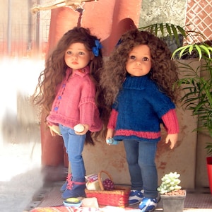 Op de afbeelding: Twee poppen met lang krullend haar dragen gebreide truien en jeans. De pop links draagt een roze trui met een blauwe knoop en een blauwe strik in haar haar. De pop rechts draagt een blauwe trui met een roze rand en een blauwe strik in haar haar. Ze staan voor een houten constructie met een picknickmand en een groene plant.