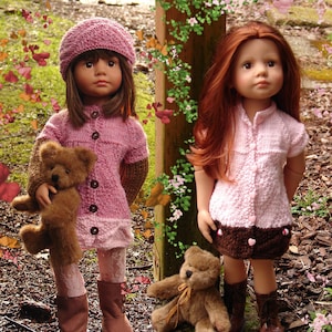 Puede incluir: Dos muñecas con vestidos de punto rosa y botas marrones sostienen osos de peluche. Las muñecas tienen el pelo largo y están de pie frente a un poste de madera con enredaderas verdes.