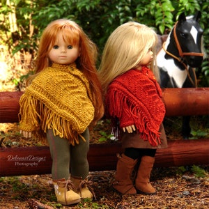 May include: Two dolls wearing colorful knitted ponchos and boots stand in front of a wooden fence. The doll on the left is wearing a yellow poncho with fringe and the doll on the right is wearing a red poncho with fringe.