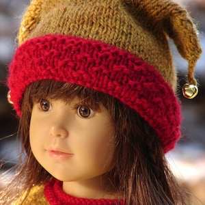 May include: A doll wearing a knitted hat with a red and brown color scheme. The hat has a pointed top and a bell on each side. The doll is wearing a matching sweater.