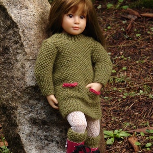 Op de afbeelding: Een pop die een groene gebreide trui draagt met rode bies en witte kanten panty's. De pop heeft bruin haar en blauwe ogen en staat naast een grote grijze rots.