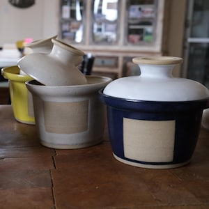 Può includere: Una collezione di burriere in ceramica con coperchi. I piatti sono bianchi, gialli e blu navy, con un'etichetta quadrata color naturale sul davanti. I coperchi sono bianchi con un bordo color naturale. Su una superficie di legno.