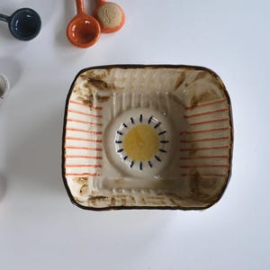Puede incluir: Un conjunto de cucharas de servir de cerámica y un cuenco cuadrado. Las cucharas son de varios colores, incluyendo amarillo, marrón y naranja, con mangos de madera. El cuenco tiene un diseño de sol y rayas naranjas.
