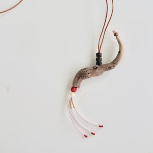 Peut inclure: Un collier en cordon de cuir brun avec un pendentif en bois en forme de branche incurvée. Le pendentif est décoré de perles noires et dorées et d'une perle rouge au bout d'un pompon.