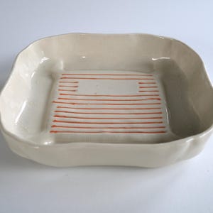 Pode incluir: Um prato de cerâmica artesanal, branco sujo, com formato quadrado e bordas suavemente curvas. O interior apresenta um design retangular com linhas horizontais laranja. Este prato decorativo é adequado para servir ou expor.