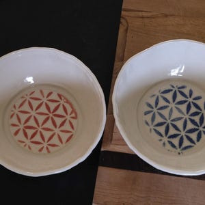 May include: Two white ceramic bowls with a floral pattern in the centre. One bowl has a red pattern, and the other has a blue pattern. The bowls are on a wooden surface and a black surface.