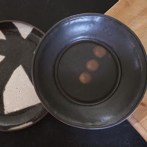 Peut inclure: Deux assiettes en céramique. L'une est noire et blanche avec un motif géométrique. L'autre est gris foncé avec trois taches de couleur cuivre au centre. Les assiettes sont sur une surface sombre avec une planche à découper en bois en arrière-plan.