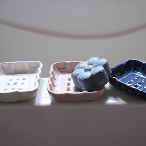 May include: Three ceramic soap dishes in white, pink, and blue. The dishes have a textured surface and are designed to hold soap bars. A blue soap bar is sitting in the pink dish.