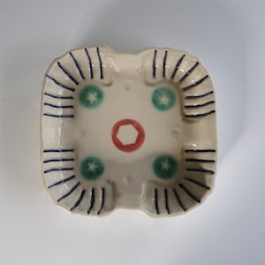 Puede incluir: Cenicero cuadrado de cerámica con base color crema. El cenicero presenta un círculo rojo en el centro, tres diseños de estrella verde y detalles a rayas azules. El cenicero tiene cuatro muescas para apoyar cigarrillos.