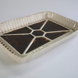 Puede incluir: Bandeja rectangular de cerámica color crema con un diseño geométrico marrón. La bandeja tiene un borde elevado con líneas verticales y detalles punteados. Ideal para servir o exhibir, mide unos 25 cm de largo.