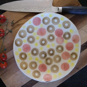Könnte beinhalten: Ein runder weißer Keramikt weißer Keramikteller mit einem wiederkehrenden Muster aus braunen, texturierten Ringen, soliden rosa Kreisen und kleinen gelben Punkten. Ein silbernes Kochmesser mit gehämmertem Finish und dunklem Griff liegt neben einer Tomatenrispe.